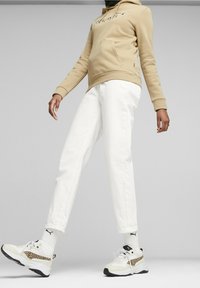 Sudadera beige con bolsillo frontal y logo, pantalones de mezclilla blancos, zapatillas blancas con acentos de estampado de leopardo y suelas negras.