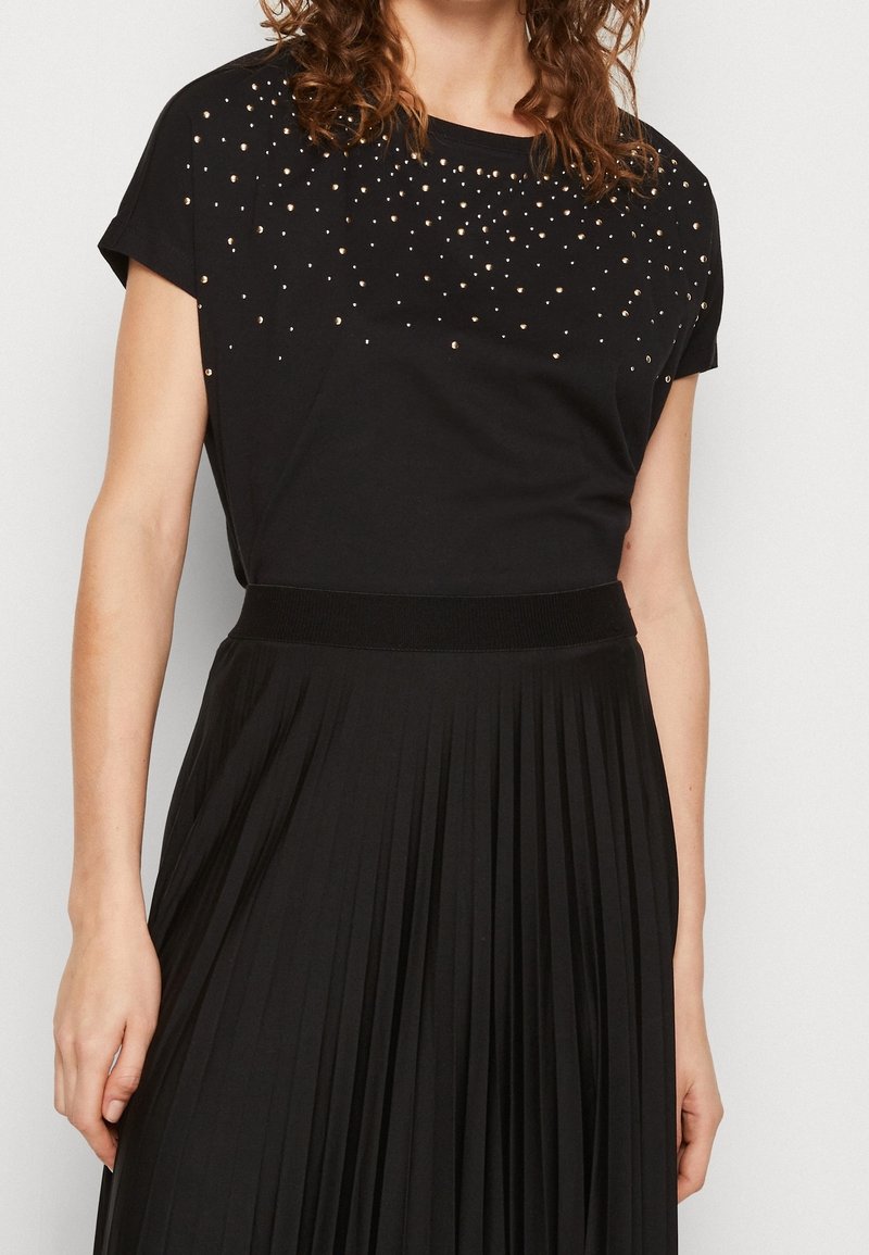 Femme portant un haut noir à manches courtes avec des clous dorés et une jupe plissée noire taille haute sur un fond clair.