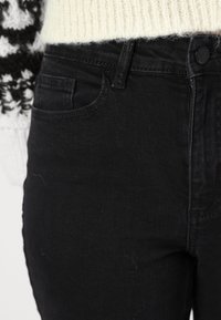 Melnas džinsas ar pieguļošu dizainu. Satur priekšējo kabatu, standartizētu pogu aizdari un faktūru ar nelielu elastību.