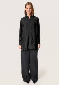 Camisa negra de manga larga con cierre de botones en el frente y un corte holgado, combinada con pantalones negros de pierna ancha y zapatos de punta. Textura suave.