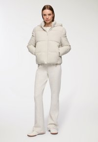 Młoda kobieta stoi ubrana w jasnobeżową kurtkę puchową z kapturem, pasujące szerokie spodnie i białe tenisówki na białym tle.