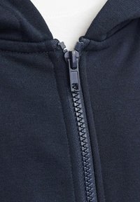 Dettaglio di una felpa con zip di colore blu navy, caratterizzata da una zip liscia e un tessuto elastico. La trama è a costine e la vestibilità è rilassata attorno al collo.