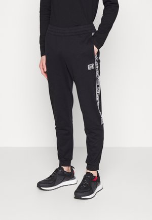 Personne portant un pantalon de jogging noir EA7 Emporio Armani avec des bandes à logo, des baskets noires avec semelles blanches, debout avec une main dans la poche.