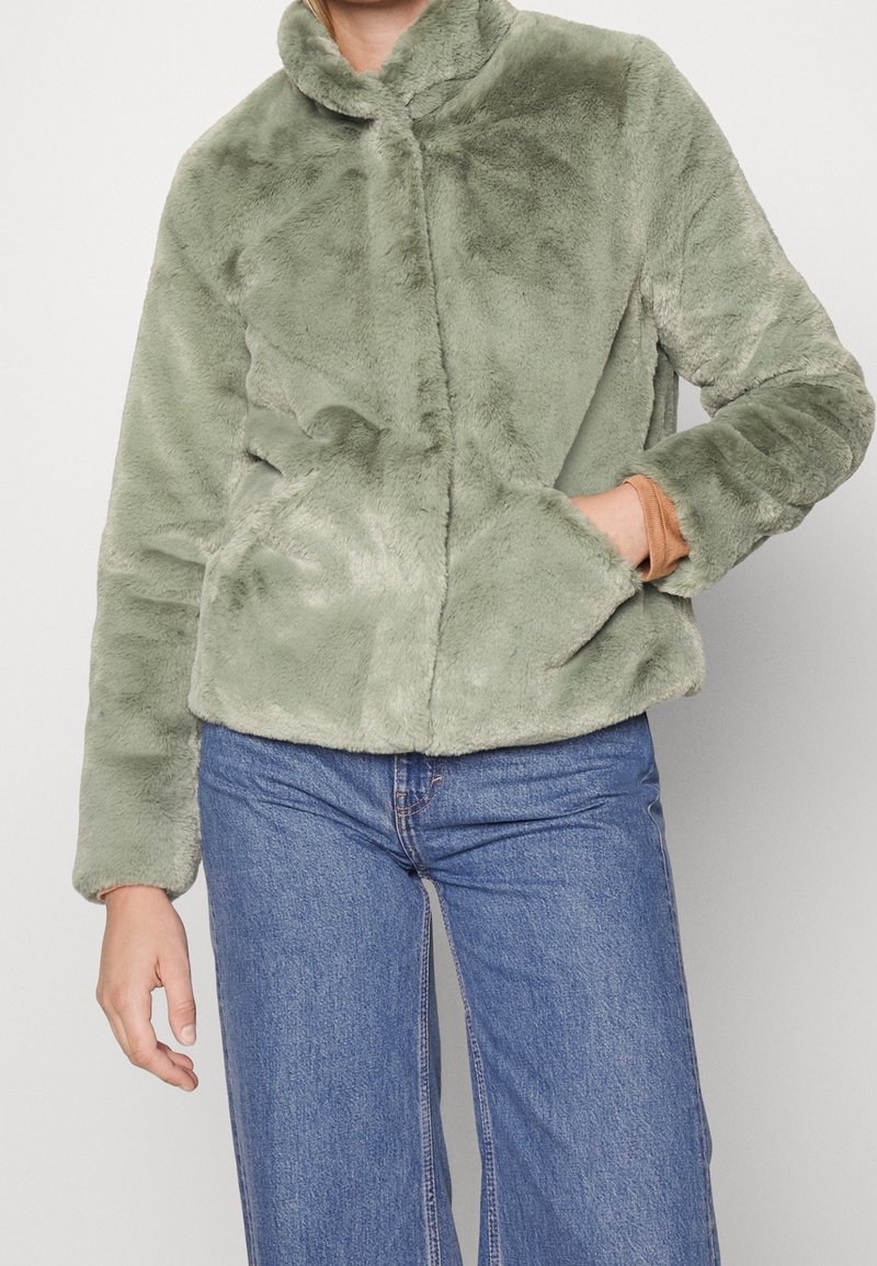 Personne portant une veste en fausse fourrure peluche vert clair avec un col montant et un jean bleu, une main dans la poche de la veste, sur fond uni.