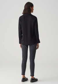 Camicia nera a maniche lunghe, abbinata a pantaloni slim-fit grigio scuro e scarpe nere, vista dal retro su uno sfondo neutro.