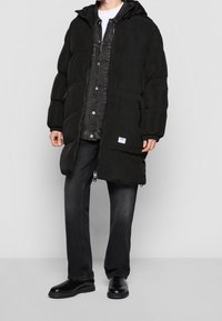 Sort dunjakke med hætte, der har lynlås foran, to sidelommer og elastiske manchetter. Båret over en mørk denimskjorte og sorte bukser.