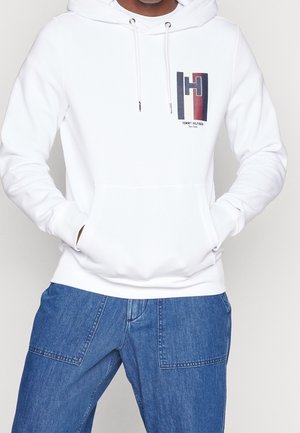 Sudadera blanca con un bolsillo frontal, capucha con cordón y un logotipo impreso con acentos en rojo y azul. Se lleva con pantalones de mezclilla azul.
