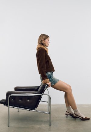 Giacca marrone corta con colletto in pelliccia sintetica, pantaloni shorts in denim e scarpe con tacco marroni. La sedia è in pelle nera con una base in cromo e bottoni.