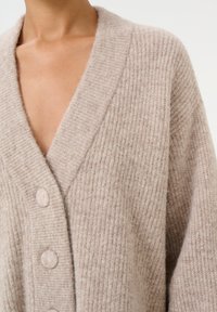 Béžový pletený svetr s žebrovanou strukturou, hlubokým výstřihem do V a velkými kulatými knoflíky. Měkký materiál s mírně chlupatým povrchem.