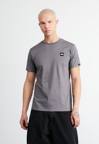 Graues T-Shirt mit kurzen Ärmeln aus Baumwolle, das ein kleines Logo-Patch auf der Brust und ein kontrastierendes Etikett am Ärmel aufweist.