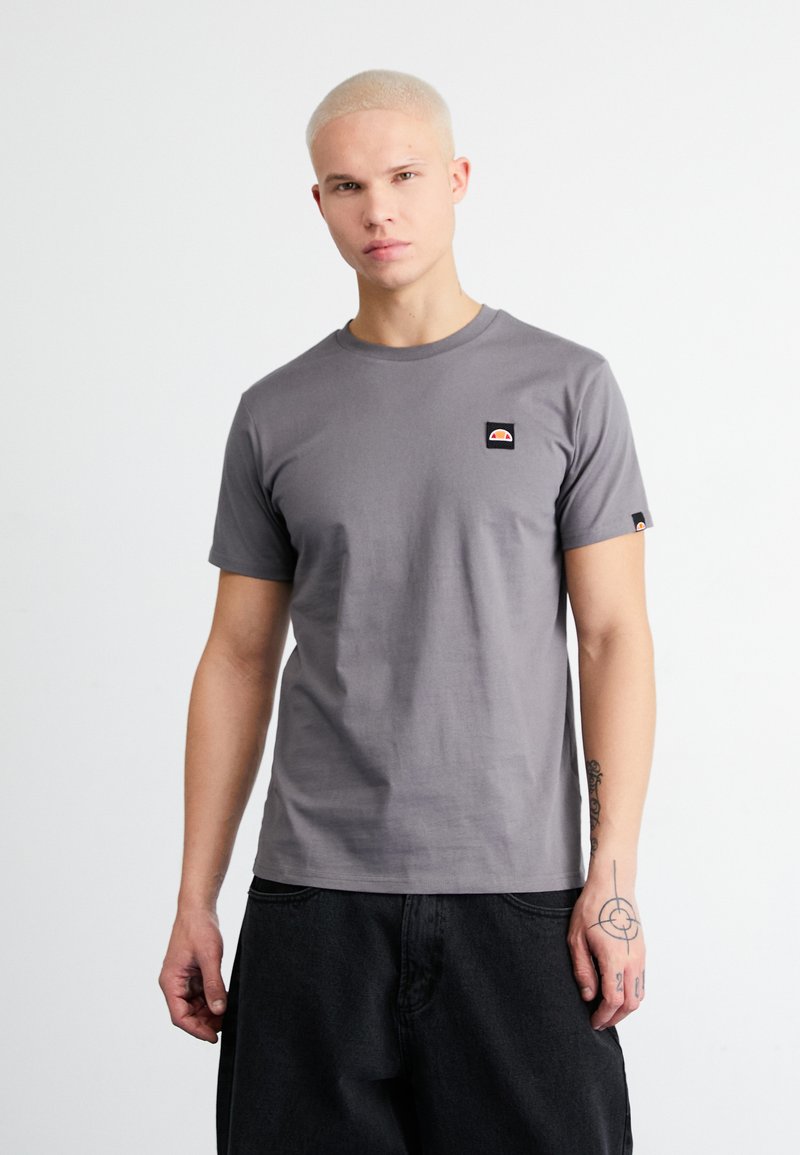 Graues T-Shirt mit kurzen Ärmeln aus Baumwolle, das ein kleines Logo-Patch auf der Brust und ein kontrastierendes Etikett am Ärmel aufweist.