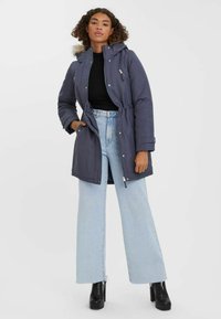 Temno modra parka s kapuco obrobljeno s krznom, zategnjenjem v pasu in sprednjim zadrgo, v kombinaciji s svetlo modrimi širokimi kavbojkami in črnimi škornji s peto.