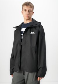 Mladic nosi crnu jaknu Helly Hansen s kapuljačom, mornarsko plavu majicu s grafikom i sive hlače na jednobojnoj bijeloj pozadini.