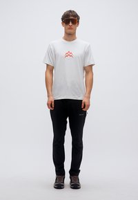 Λευκό μπλουζάκι με κόκκινο γραφικό βουνού, μαύρα παντελόνια και καφέ παπούτσια πεζοπορίας. Το μοντέλο φοράει oversized γυαλιά ηλίου με φακούς πορτοκαλί απόχρωσης.