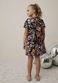 Schwarz-blumiges Kleid mit mehrfarbigen Pailletten, kurzen Ärmeln und einem Schlüssel-ausschnitt hinten. Kombiniert mit glitzernden, pinken Schuhen. Silbergraue Discokugeln in der Nähe.