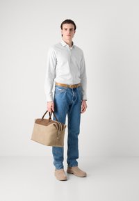 Beige Reisetasche mit braunen Akzenten, strukturiertem Material und doppelten Griffen. Das Modell trägt ein hell gestreiftes Hemd, blaue Jeans und beige Schuhe.