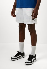 Hvite treningsshorts med subtilt mønster, kombinert med svart-hvite joggesko. Modellen har på seg hvite ribbestrikkede sokker og en blå T-skjorte.