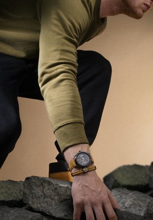 Orologio da uomo con quadrante nero, dettagli argentati e cinturino in pelle beige. Un braccialetto intrecciato con un ciondolo completa l'outfit. Manica verde oliva visibile.