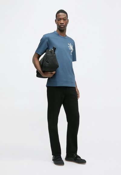 T-shirt azul de manga curta com desenho floral branco, combinado com calças pretas e uma bolsa preta. A modelo está de pé de forma relaxada com uma expressão neutra.