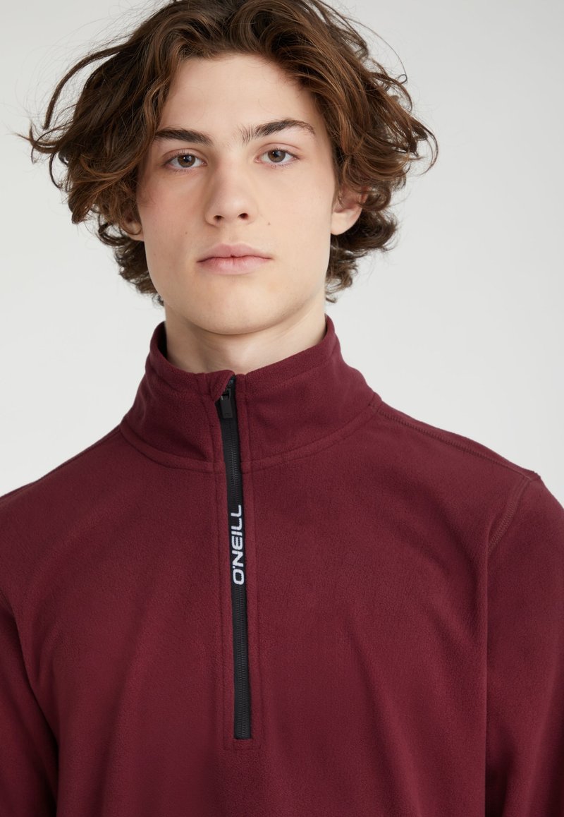 Pull-over en polaire bordeaux avec zip noir arborant un logo de marque. Texture douce et design à col haut.