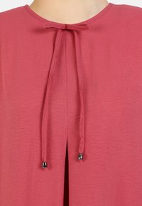 Rosa top med rund halsudskæring, en frontslids og en sløjfedetalje med slanke bånd, der ender i metalliske spidser. Struktureret stof.