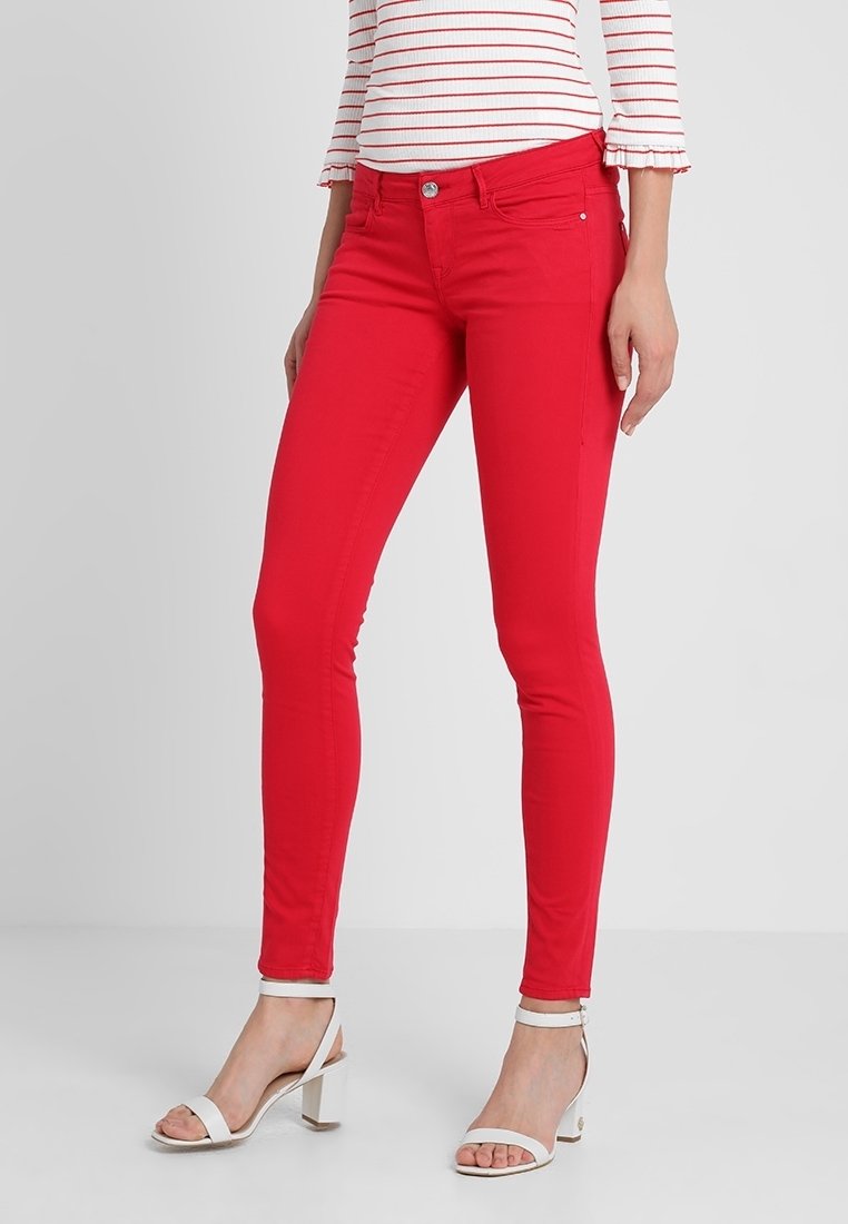 Jean skinny rouge en denim, avec une fermeture à boutons sur le devant et cinq poches. Porté avec un haut à rayures et des sandales blanches à fines lanières.