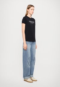 Svart bomull T-skjorte med "Armani Exchange" logo, kombinert med lyseblå vid ben denimjeans med et trykt mønster, og beige sneakers.