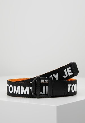 Czarny materiałowy pasek z grubym białym napisem „TOMMY JEANS”, pomarańczowym spodem i czarną metalową klamrą, pokazany zwinięty na białej powierzchni.