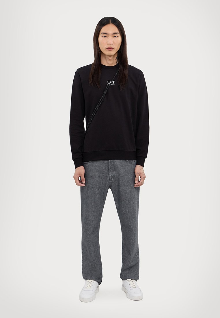 Sweatshirt noir avec logo, jean droit gris, et baskets blanches. Cheveux longs et droits, se tenant devant un fond uni.