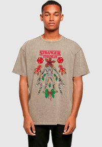 Hnědý bavlněný tričko s barevným grafikou „Stranger Things“, zobrazující postavu obklopenou prvky s vánoční tématikou a cukrovými tyčinkami.