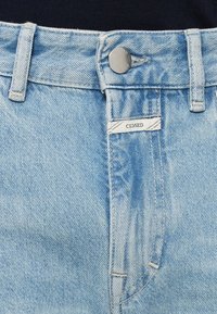 Ceinture de jeans en denim bleu clair avec bouton argenté, passants pour ceinture et une étiquette blanche indiquant « CLOSED » au-dessus de la braguette.