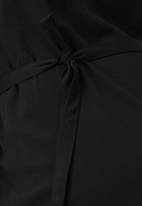 Abito da maternità nero con nodo frontale, texture liscia e vestibilità ampia, progettato per accogliere una pancia in crescita.