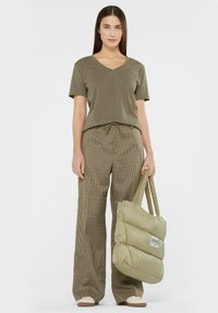 Olive green V-neck t-shirt, tan and brown plaid high-waisted pants, and a puffy green tote bag with a logo, worn with beige slip-on shoes.