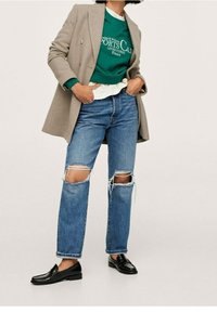 Lichtgrijze blazer over een groene sweater, blauwe gescheurde jeans en zwarte loafers. Kenmerken zijn onder andere een dubbelzijdig ontwerp en ruwe denimranden.