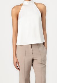 Haut blanc à col halter en tissu texturé, avec un col montant et une coupe fluide, accompagné de pantalons beiges ajustés.