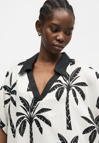 Femme aux cheveux tressés portant des boucles d'oreilles créoles en or et un chemisier blanc avec des imprimés de palmiers noirs et un col noir, regardant vers le bas.