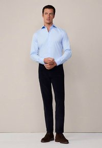 Ljusblå bomullsskjorta med spetsig krage, långa ärmar och knappstängning, tillsammans med mörka byxor och bruna skor.