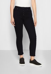 Jeans skinny azul escuro com fecho de cinco botões, bolsos frontais e uma textura suave, combinados com sapatilhas pretas de cano baixo.