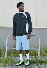 Sort og hvid atletisk langærmet skjorte, blå denimshorts og grønne og sorte sneakers med en kraftig sål og sorte snørebånd.