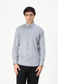 Chemise à manches longues à boutons en tissu blanc avec un motif géométrique bleu, col boutonné et boutons foncés, associée à un pantalon sombre.