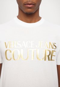 Balta medvilnės T-marškiniai su ryškiu auksiniu folijos spaudiniu, kuriame užrašyta "VERSACE JEANS COUTURE" didelėmis, didžiosiomis raidėmis.