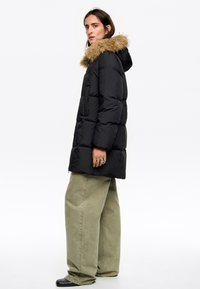 Czarna puchowa kurtka z kapturem obszytym futrem, posiadająca pikowany wzór, frontowy zamek błyskawiczny i boczne kieszenie, zestawiona z oliwkowymi spodniami o szerokich nogawkach.