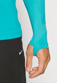 Turkos långärmad träningsoberdel i stretchmaterial, med texturerade mönster på ärmarna, svart midjeband och en Nike-logotyp.