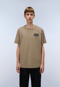 Khaki tričko vyrobené z bavlny, má krátké rukávy, kulatý výstřih a černou nášivku s logem na levé části hrudi.