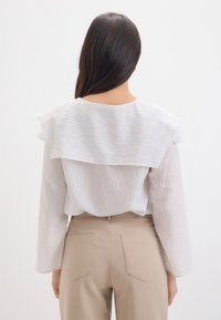 Kvinde med langt mørkt hår iført en hvid skjorte med lange ærmer og stor krave samt beige bukser med høj talje, set fra bag.