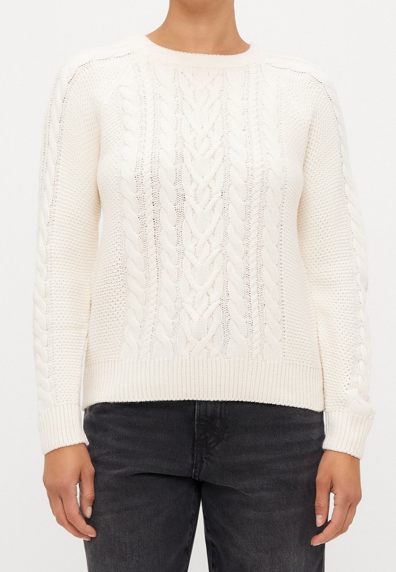 Personne portant un pull en maille torsadée blanc avec des manches longues et des poignets côtelés, associé à un jean en denim noir.