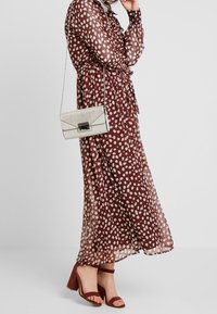 Hnědá maxi šaty s puntíky z průhledné tkaniny s páskem na zavázání, doplněné vínovými sandály na podpatku a krémovou kabelkou přes rameno se stříbrnými detaily.