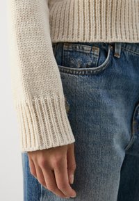 Kremfarget strikkegenser med ribbestrikkede mansjetter, kombinert med blå slitte denimjeans. Hånden hviler synlig mot livet.