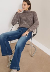 Suéter marrón de cuello alto de punto, pantalones anchos de mezclilla azul, sentado en una silla cromada, alfombra texturizada de color claro debajo.