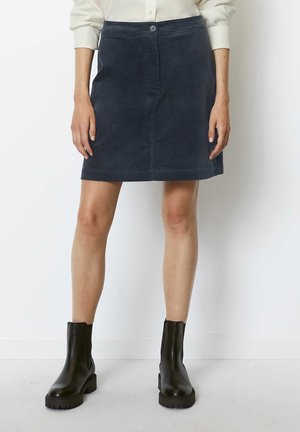 Frau in einem knielangen, marineblauen Rock, cremefarbenem Langarmshirt und schwarzen Stiefeletten, die vor einem weißen Hintergrund steht.
